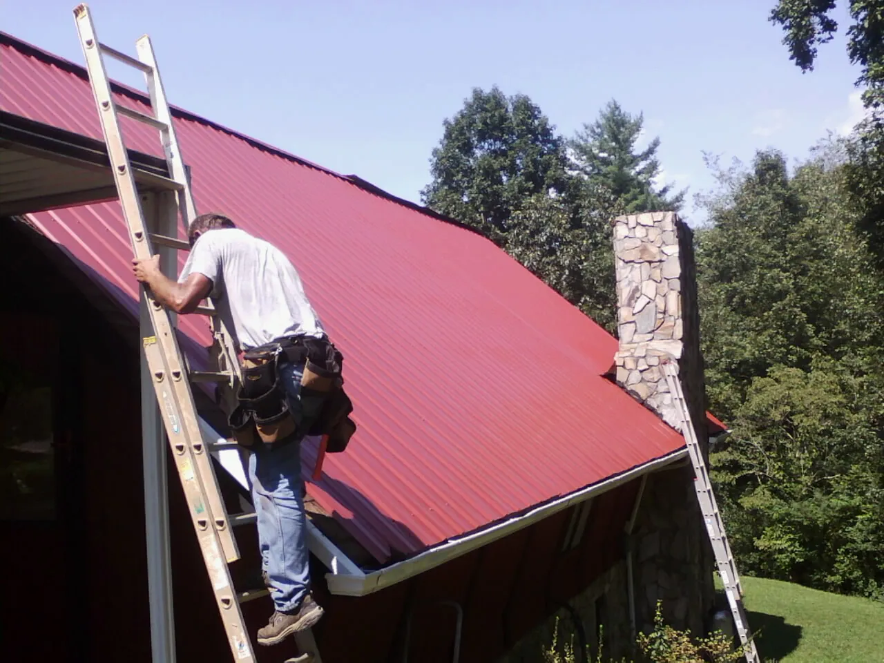 Roofer on a ladder beside a red metal roof and stone chimney — Soffit & Fascia Repair by Roofing Brothers in Bonnieville